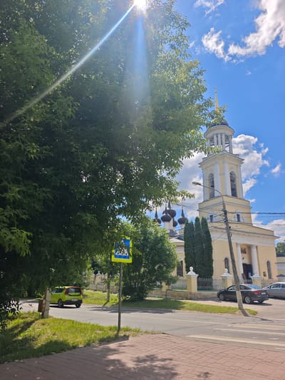 This is a full shot of a street scene in Chekhov, Russia, on a sunny day. A large, leafy green tree dominates the left side of the frame, its branches extending into the upper portion of the image where the sun's rays create a lens flare effect. In the background, a yellow church with a tall bell tower stands against a bright blue sky with scattered white clouds. The church has a dome and a golden cross at its apex. A pedestrian crossing sign is visible in the middle ground. To the left of the sign, a bright green car is parked on the side of the road. Further to the right, a dark-colored sedan is parked in front of the church. The foreground shows a brick-paved sidewalk and a section of asphalt road with painted white lines. The overall impression is a quiet, sunlit day in a town with a prominent church.