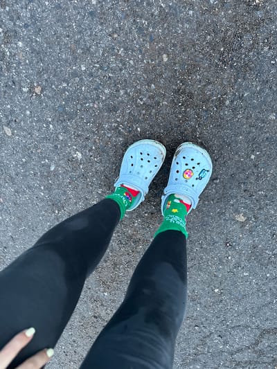 The image shows a pair of light blue Crocs with jibbitz, worn with green Christmas-themed socks and black leggings. The Crocs are on a person's feet, which are positioned on a dark asphalt surface. The asphalt appears to be rough and textured, with small stones and debris visible. The lighting suggests it is daytime. The overall scene is a close-up, overhead view of the person's lower legs and feet. The location is described as the city of Zhebrei, Russia.