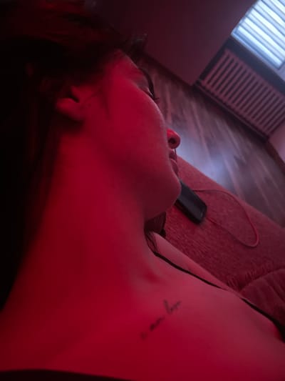 The image shows a close-up, side profile of a person lying down, illuminated by red light. The person's face is partially visible, with their eye closed and their head turned away from the camera. A tattoo is visible on their collarbone. A black object, possibly a phone, and a red cord are on the surface next to them. In the background, a tanning bed is visible. The overall scene suggests a private, indoor setting.