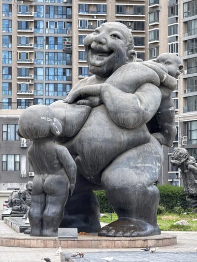 The image depicts a large, bronze-colored statue of a human figure with multiple smaller figures entwined around it. The statue is located outdoors, in an urban area with tall buildings in the background. There is a sidewalk with pigeons, and greenery. The location is in Changping District, China.