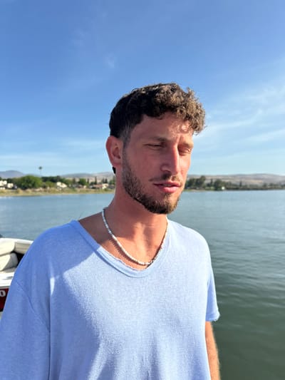 A young adult male with short, curly brown hair and a beard is wearing a light blue v-neck t-shirt and a beaded necklace. He is standing on a boat on a lake with a light blue surface, looking down and to the right, with his eyes closed. The shoreline is in the distance, with vegetation visible. The sky is blue with some cloud cover. This image was taken near the Sea of Galilee in Migdal, Israel. The image is taken in a portrait orientation.