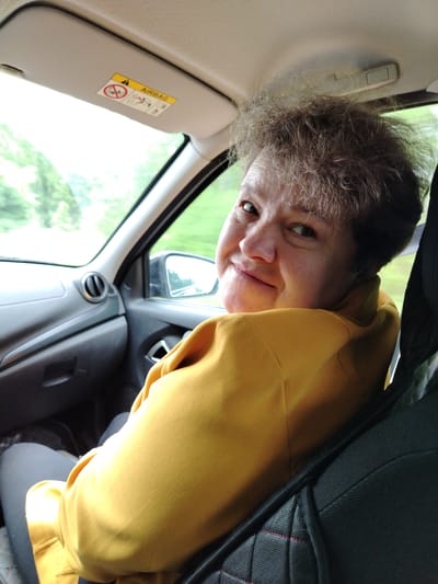 A woman in a mustard-yellow jacket is looking back over her shoulder, smiling at the camera. She is sitting in the passenger seat of a car. An "AIRBAG" warning label is visible on the sun visor. The car is moving, as evidenced by the blurry trees visible through the window. The interior of the car is grey and black. I cannot determine the specific location within the city of Краснояр, Russia, from the provided image.