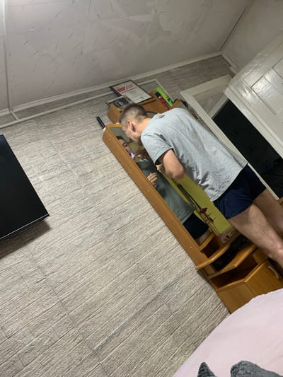 In the city of Belogorsk, Russia, a man is inspecting his teeth in a mirror. He is wearing a grey t-shirt and navy blue shorts. The man is standing in front of a wooden vanity with shelves, and the floor is covered in light-colored wood-effect tiles. To the left of the vanity, a portion of a black television screen is visible. In the background, a white door and a white ceiling with marble-patterned panels are present. The overall environment suggests an indoor domestic setting.