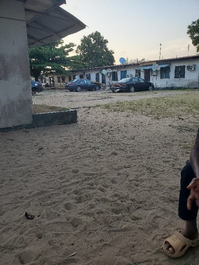 The image displays an unpaved outdoor area in Isunba, Nigeria, under a clear sky. The ground consists of sand and dirt, marked with numerous footprints and sparse dry vegetation. In the bottom right quadrant, the lower right leg and foot of a person are visible, clad in dark trousers and a tan molded sandal.

In the mid-ground, a row of single-story, light-colored buildings extends horizontally across the frame. These structures feature multiple windows, several equipped with dark coverings, and exterior-mounted air conditioning units and satellite dishes (at least five visible). The buildings have visible roofs, appearing to be corrugated metal. Three dark-colored sedans are parked in the sandy area in front of these buildings: one partially obscured on the far left, another facing right in the center, and a third facing left on the right, which shows a visible mark or scuff on its rear bumper.

A large, leafy tree is prominent in the background behind the buildings on the left. A section of a different building with a wide, overhanging roof is visible in the immediate left foreground. No overt actions or interactions are discernible beyond the static arrangement of subjects and objects within the scene.