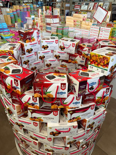 The image depicts a retail display of packaged confectionery, primarily featuring boxes of "Kestane Şekeri" (chestnut candy) and other sweets. The products are stacked in an organized manner, creating a tiered display. The packaging is predominantly red, white, and brown, with prominent branding for "Kibarzade" and images of chestnuts and chocolate. Some boxes are designed as gift bags with red rope handles. In the background, shelves are filled with more boxes of confectionery and other retail items, suggesting a store or market setting. The context provided mentions the city of Acıpayam, Turkey, which is known for its chestnut production, aligning with the prominent product being featured. The lighting appears to be artificial, typical of an indoor retail environment. No people are visible in the image.