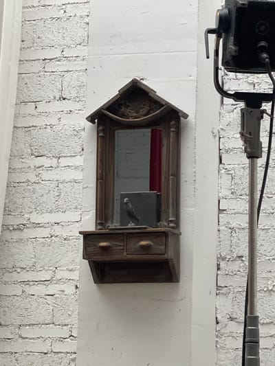 A bird figurine stands on a shelf in front of a mirror. The mirror is in a wooden frame shaped like a house with two drawers below it. The background is a white brick wall and a white column. A lighting fixture is on the right side of the image.