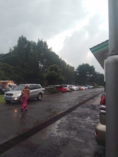 The image captures an outdoor parking area in Gitaru, Kenya, under a heavily overcast sky, suggesting either recent rain or a very cloudy day. The asphalt ground is visibly wet and features a long, dark metal drain grate running diagonally from the lower left to the center midground.
In the midground, a person wearing a striped top (red, orange, black) and a long pink or maroon skirt/wrapper with a matching head covering walks away from the camera, heading towards a silver Toyota RAV4 SUV. The SUV has its headlights illuminated, and its Kenyan license plate, "KCK 240J," is clearly visible on the front.
Further into the background, numerous other vehicles of various makes and colors, including white, red, and beige, are parked in rows. Several indistinct figures can be seen near these parked cars. The background is dominated by dense green trees and lush foliage. On the far right, a section of a building with a green-edged roof is partially visible, alongside a dark grey pole that obscures part of a beige car. On the far left in the distance, a white truck or van is discernible, with some faint, illegible text on its side. The overall scene conveys a muted, possibly damp atmosphere.