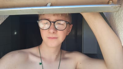 This media file shows a close-up, chest-up portrait of a young person with blonde hair and glasses. They are looking directly at the camera, with a neutral expression. The person is shirtless and wearing a green necklace with a small charm. They are framed by a wooden structure at the top and a metal bar across their forehead. The background is dark, suggesting an indoor setting, possibly with a window or doorway to the right. The lighting appears to be natural and coming from the front and slightly above, creating highlights on the person's face and skin. The image has a casual, candid feel, suggesting it might be a selfie. The context provided indicates the location is Parchim, Germany, but there are no specific visual cues in the image to confirm this. The time of day is not explicitly indicated, but the lighting suggests it could be daytime. There is no visible text in the image.