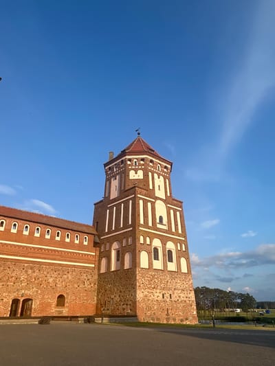 The image depicts a tall, octagonal brick tower with a red tiled roof, part of a larger red brick castle complex. The tower has a weathered stone base and is decorated with white arched windows and vertical white rectangular accents. A long, low wing of the castle, also constructed of red brick with a red tiled roof, extends to the left, featuring a series of evenly spaced white arched windows. The castle is set against a bright blue sky with wispy white clouds and some distant trees. In the foreground, there is a paved area, likely a courtyard or pathway, and the edge of a grassy area with some distant vehicles. The lighting suggests it is either early morning or late afternoon, with long shadows cast on the ground. This scene is located at the Mir Castle Complex in the village of Mir, within the Zhukhavitski Sel'ski Savet of Belarus.