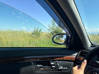 This media file captures a POV perspective from inside a car, looking out the partially open window on a sunny day. The scene is a rural landscape with tall, dry grass and scattered trees under a clear blue sky. The interior of the car features a dark dashboard with wood grain accents and chrome detailing on the door panel, including the door handle and a set of control buttons. A side mirror is visible, reflecting a portion of the sky and trees. A woman's hand, with neatly manicured nails, rests on the steering wheel, indicating that the car is in motion or about to be. The overall impression is one of a peaceful, scenic drive on a warm day in Ivanyane, Bulgaria. There is no visible text in the image.