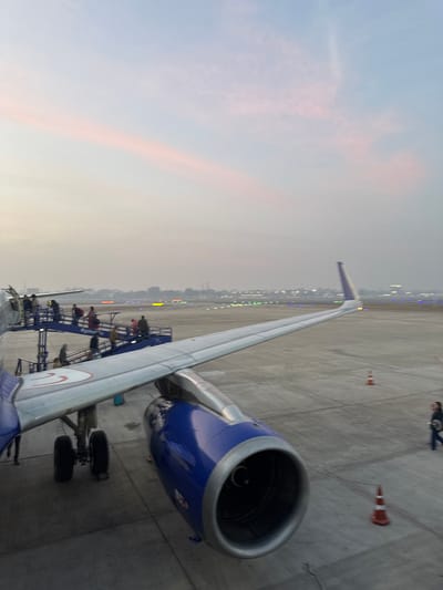 An airplane stands on the tarmac during twilight. Passengers are ascending a mobile staircase to board. The plane's wing extends towards the lights of a distant city. Dominating the right side is a blue and silver jet engine. Orange traffic cones are nearby. The sky is filled with pale colors, hinting at either sunrise or sunset. A figure walks in the distance.