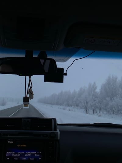 View from inside a car driving on a snow-covered road. Trees covered in frost line the road in the distance, under a gray sky. A dashboard camera and air freshener are mounted on the rearview mirror. The car radio displays a list of FM radio stations including Yuldash, Sputnik, Shanson, and Like FM, and a station at 101.80 MHz.
