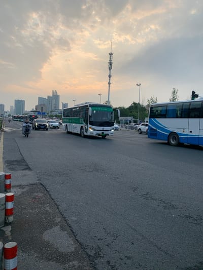 The image shows a street scene in Wuhan, China, located at the coordinates 30.51 latitude and 114.404 longitude. The scene is captured during the day, as evidenced by the overcast sky, with some sunlight breaking through the clouds. Two buses are parked on the street, one is white with green trim and the other is blue and white. There are several cars driving on the street. A man on a motorcycle is riding along the street on the left side. The street is asphalt and there are red and white cylindrical traffic barriers on the left side of the image. The street is lined with buildings on both sides. Some of the buildings are tall and modern. In the background, there is a tall, slender radio tower, which stands out against the sky. The scene is peaceful and there is a sense of normalcy.