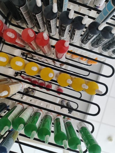 The image displays an organized collection of liquid pigment dispensers within the Municipal Unit of Acharnes, Greece. Numerous transparent, cylindrical tubes, each equipped with a white plastic nozzle, stand vertically in parallel rows. These tubes are supported by a multi-tiered, black wire rack featuring S-shaped horizontal supports. The pigments inside are visibly colored in distinct rows, predominantly red, yellow, green, and dark grey/black, with additional tubes containing orange and light brown hues. The arrangement suggests a systematic storage or retail display. The background consists of a plain, light-colored wall, with a portion of a white tiled floor discernible in the lower right corner. No individuals or specific activities are depicted.