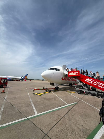 The image depicts a Jet2.com passenger airplane at an airport, likely in Uttlesford, United Kingdom. Passengers are disembarking using a set of mobile stairs bearing the Jet2.com logo. The aircraft has a white fuselage with red and yellow Jet2.com branding. The sky is partly cloudy. Several ground vehicles are visible on the tarmac. Another airplane, partly visible in the distance, has a tail fin with a blue and yellow design. The ground is concrete with painted white and green lines delineating parking and movement areas.