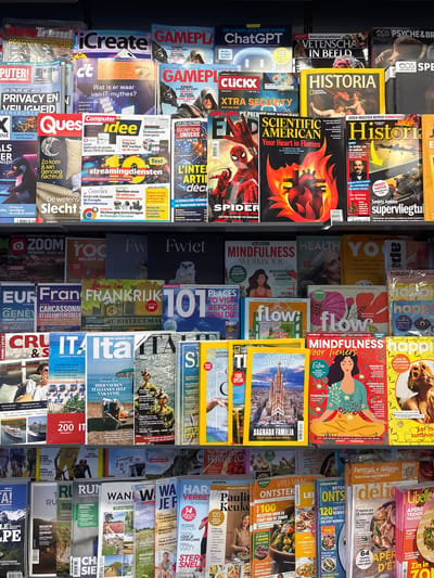 The image displays a large collection of magazines stacked vertically. The magazines feature a variety of subjects, including science, history, technology, and lifestyle. Many of the covers depict people, often in posed or candid shots, engaged in different activities. Some magazines have illustrations or graphics as their main focus, such as the fiery imagery on one cover or abstract designs on others.

Several magazines are identifiable by their prominent titles. These include "Historia," "Scientific American," "Game Play," "Create," "Vrouwen," and "National Geographic." The text on the covers is in various languages, primarily Dutch and English, with some French also visible. Words like "Historia," "Scientific American," "Game Play," "Create," "Vrouwen" (meaning women), and "National Geographic" are legible.

The setting appears to be a retail environment, possibly a magazine store or a display rack. The lighting is somewhat dim, suggesting an indoor location. There are no clear indicators of time of day or weather. The overall impression is one of a diverse selection of reading material, catering to a range of interests. The magazines are neatly arranged, suggesting a well-maintained display. The image is a snapshot of a typical magazine stand, showcasing the variety of content available to consumers.
