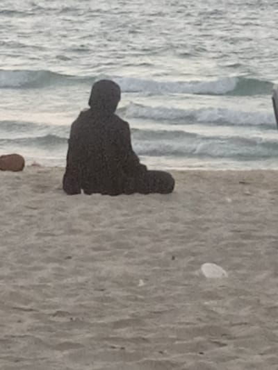 An individual clad in a dark, full-body garment, including a head covering, is seated facing the sea on a sandy beach in Al-Mawasi, Palestinian Territories. The person is positioned with their back to the camera, appearing to sit cross-legged or with bent legs. To the left of the individual on the sand, a brown, organic-looking object rests. The foreground beach consists of light-colored sand, upon which a small, white, translucent object is visible towards the lower right. The background features the sea with gentle waves, exhibiting light blue and grey tones. The scene is illuminated by diffused light.