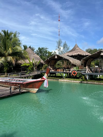 The image depicts a scenic view of a waterfront setting with traditional Thai architecture and a longtail boat. The scene is dominated by a large body of calm, turquoise water, likely a river or bay, with ripples suggesting a gentle current or a fountain in the background. Adjacent to the water is a wooden dock leading to a longtail boat, its prow adorned with colorful fabric streamers.
Behind the dock, several huts with conical, thatched roofs are visible, interspersed with lush green tropical foliage, including palm trees. The structures appear to be part of a resort or restaurant, designed in an open-air style. A life preserver ring hangs from a post near one of the huts. In the distance, a tall, red and white telecommunications tower pierces the bright blue sky, which is dotted with wispy white clouds.
The overall impression is one of a sunny day in a tropical location. The light suggests it's daytime, likely mid-morning or afternoon. The vibrant colors of the water, greenery, and boat, along with the clear sky, evoke a peaceful and inviting atmosphere. There are no people visible in the image, focusing the viewer's attention on the natural beauty and architectural elements of the place. The location cues, such as the tropical vegetation and the longtail boat, combined with the provided location context of Tambon Pa Kae Bo Hin, Thailand, strongly suggest the setting is in Thailand. There is no visible text in the image.