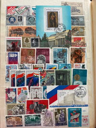 The image is a close-up overhead view of a page from a stamp album filled with postage stamps from the Soviet Union and other countries. The stamps depict various themes including historical events, art, architecture, and space exploration.

One stamp, located in the upper center of the image, features a depiction of the Moscow State University, a prominent landmark in the city of Moscow, Russia. The stamp, labeled "ПОЧТА СССР 1991", also shows a postmark with "МОСКВА - ПОЧТАМТ" indicating its origin. While the university is located in Moscow, there is no indication in the image that this location is related to the city of Сокол, Russia.

The page is organized with stamps arranged in rows and columns, some overlapping. The background of the album page appears to be a light-colored paper, possibly off-white or cream. The lighting suggests an indoor setting with diffuse light. No people are visible in the scene, and the focus is entirely on the collection of stamps. The stamps themselves vary in color, size, and design, showcasing a diverse range of imagery.