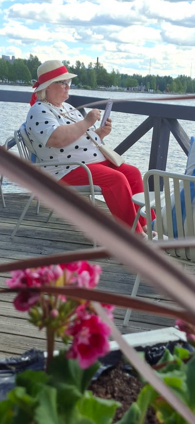 A woman wearing a white polka dot shirt, red pants, and a straw hat with a red ribbon is sitting on a chair on a wooden deck next to a body of water. She is looking at her phone, which she holds in her hands. A small, cream-colored clutch bag with a gold chain strap rests on her lap. In the foreground, out of focus, are pink flowers in a planter. The background shows a tree-lined shore and a cloudy sky. The scene appears to be during the daytime, possibly late spring or summer, given the lush greenery and the woman's attire. The location is identified as Lahti, Finland.