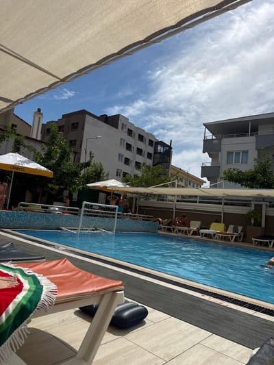 The image depicts an outdoor swimming pool area, likely a hotel or resort, with people lounging on chairs and swimming. The pool is rectangular and filled with blue water. A white water polo goal is set up in the shallow end of the pool. Several white lounge chairs with colorful towels are arranged around the poolside. In the background, there are multi-story buildings with balconies and trees. A large, light-colored awning stretches over part of the seating area, providing shade. The sky is partly cloudy and blue. The presence of lounge chairs, umbrellas, and a well-maintained pool suggests a recreational setting. Given the context of Ayvalık, Turkey, this scene likely represents a tourist accommodation.