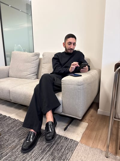 A male individual is seated on a light gray two-seater sofa in an indoor setting in Yaqum, Israel. The man, with short dark hair and a beard, is dressed in a black long-sleeved shirt, black wide-leg trousers, and black patent leather loafers. He holds a black smartphone in his left hand and interacts with its screen using his right index finger. A second black smartphone lies face down on the sofa cushion to his right. The floor is covered by a patterned area rug in shades of gray, black, and off-white, with wood flooring visible on the right. A plain white wall forms the background, alongside a large, reflective glass partition on the left that shows a blurred interior reflection. A silver-colored metal furniture leg is visible on the far right.