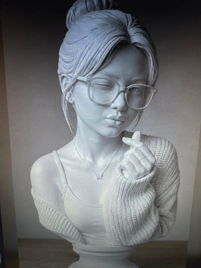 This is a digital rendering of a bust of a young woman with a topknot hairstyle and glasses. She is winking and puckering her lips, with one hand forming a heart shape gesture. She is wearing a tank top and a knitted sweater draped over her shoulders. A delicate necklace with a butterfly pendant is visible. The bust is displayed on a pedestal against a blurred, gradient background. The scene is not located in Protasovo, Russia; it is a digital artwork.
