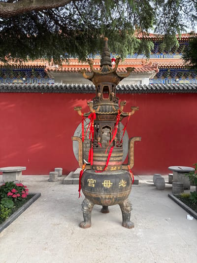 The image shows a large, elaborate bronze incense burner in an outdoor setting. The burner is decorated with intricate designs, including small dragon figures and bands of ornate patterns. Red ribbons are tied around the burner. Chinese characters are visible on the side. The burner sits on a stone-tiled area near a red wall with traditional Chinese architectural details. A tree is visible on the left side of the image and a flower bed with red flowers is visible on the bottom right. The picture was taken in Lintong District, China.