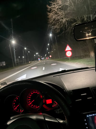 This image captures a nocturnal scene from the driver's perspective inside a vehicle traveling on a road in 23 August, Romania. The foreground features a black steering wheel, dashboard, and the car's instrument cluster, which is illuminated with red backlighting. The speedometer needle indicates approximately 60 km/h, while the tachometer shows about 2000 RPM. An odometer displays a reading of 305139 km. A section of an infotainment screen is visible on the lower right dashboard. The windshield exhibits numerous water droplets or streaks, causing visible light sources to appear diffused with radial reflections.

Through the windshield, a multi-lane asphalt road extends into the distance, illuminated by multiple streetlights mounted on poles. White dashed and solid lane markings are visible, along with a large white arrow on the road surface indicating a forward or slightly rightward direction. On the right side of the road, a signpost displays two stacked traffic signs: an upper triangular warning sign with a red border and a white background featuring three black arrows forming a circular pattern, indicating an upcoming roundabout; beneath it, a circular regulatory sign with a red border and a white background displaying the number "50", signifying a 50 km/h speed limit. Sparse, bare tree branches are discernible against the dark sky on the right side of the road. Distant red and white vehicle lights are visible further down the road. No individuals are present in the depicted scene.