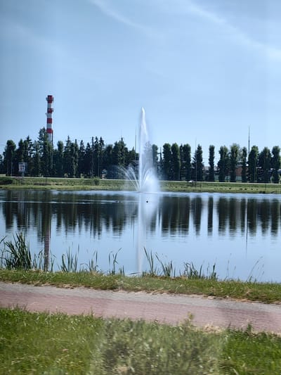 The image shows a lake with a fountain in the center, located in the city of Dzyarzhynsk, Belarus. In the background, a red and white striped smokestack is visible, along with a line of trees and some buildings. The sky is clear and blue, and the water reflects the surrounding scenery. A paved path runs along the edge of the lake in the foreground, bordered by grass. There are no people or animals visible in the frame.