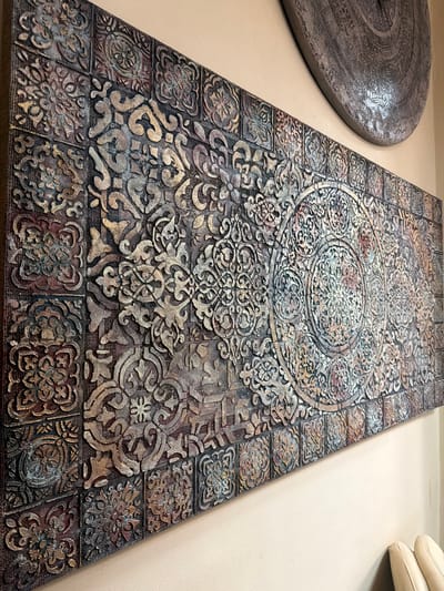 This is a close-up, eye-level, horizontal shot of a textured wall art piece with intricate, embossed patterns, suggestive of Islamic or Moroccan tilework. The artwork is rectangular and appears to be mounted on a wall. It features a repeating grid of smaller square motifs surrounding a larger central circular design. The colors are muted, with tones of deep red, gold, brown, and grey, giving it an antique or distressed appearance. The surface texture is rough and layered. In the upper right background, a portion of a large, circular, metallic decorative plate is visible, also with embossed patterns. The wall behind the artwork is a neutral beige color. The scene is set indoors, and no specific location in Maloyaroslavets, Russia can be determined from the image. There are no people, actions, or interactions depicted.