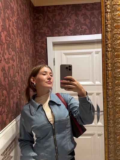 A young woman with blonde hair pulled back in a ponytail, wearing a denim jacket over a cream-colored turtleneck and a crossbody bag, takes a selfie in a mirror. She is in a room with red floral wallpaper and a white door behind her. The mirror has an ornate gold frame. The location is identified as Shkotovo, Russia.