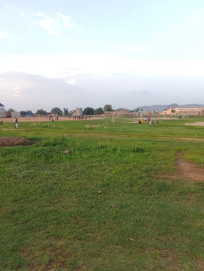 The scene depicts a grassy field in Ibo, Nigeria. In the foreground, the grass is green and somewhat overgrown, with patches of bare earth visible in the lower right. In the mid-ground, people are engaged in various activities, including what appears to be a volleyball game and other undefined sporting actions. Several buildings and structures are visible on the horizon, including a stone tower and some residential or commercial buildings. A hill is present in the far background. The sky is overcast.