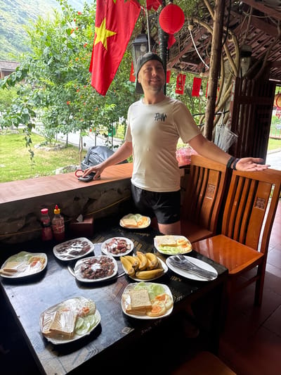 The image captures a scene in Mèo Vạc Commune, Vietnam. A man stands beside a wooden table laden with various dishes, some covered in plastic wrap. The meals include sandwiches, fruit, and meats. Utensils are also visible. He wears a light-colored shirt, dark shorts, and a cap and holds his hand up. A Vietnamese flag hangs near a dense green plant with red flowers. In the background, there is a glimpse of the surrounding landscape and structures, including wooden furniture. The setting seems to be an outdoor covered area.
