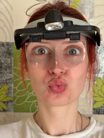 A person with reddish hair and wearing a head-mounted magnifier with a light is looking at the camera. The person is wearing a white shirt. The background is a patterned surface with floral designs. The image does not provide any information to confirm if the location is Заболотово, Russia.