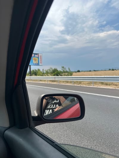 This media file shows a view from inside a car, looking out the side window and into the side mirror. The scene appears to be a rural highway in Bulgaria, likely Malo Konare, given the context. The sky is bright and cloudy, suggesting daytime with fair weather.
In the side mirror, a person wearing a black t-shirt with white text is visible. The text on the shirt is partially obscured but appears to be in all caps, with some letters like "V", "M", and "R" discernible. The person's arm and shoulder are also visible. The side mirror itself is black with a red accent on the car's body reflected in it.
Outside the car, the highway stretches into the distance, bordered by a metal guardrail and dry, grassy fields. Green trees and bushes are scattered along the roadside. In the background, a billboard is partially visible, featuring a colorful advertisement with what looks like a depiction of food, possibly a burger, and text, one part of which reads "BURGER+". The overall impression is a snapshot of a journey on a sunny day along a highway, with elements of advertising and personal reflection in the mirror.