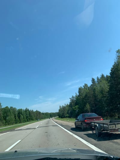 The image shows a view from inside a vehicle traveling on a paved two-lane road, likely a highway, with white lane markings. The road curves gently to the right, bordered by a grassy shoulder and a dense forest of green trees on both sides. The sky is clear blue with a few wispy clouds. On the right side of the road, a dark red Peugeot sedan is pulling a small utility trailer. Further ahead on the road, other vehicles can be seen in the distance. The scene appears to be set in a rural or semi-rural area in Belarus, given the mention of Гацукоўскі сельскі Савет, which is a rural council in the Slutsk District of Minsk Region, Belarus. The weather is bright and sunny.