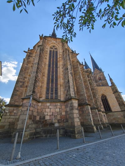 This image captures the imposing Gothic architecture of a church, likely St. Lawrence's Church in Vysoké Mýto, Czechia. The perspective is a low-angle shot, emphasizing the grandeur of the stone structure against a clear blue sky dotted with fluffy white clouds. Green leaves from an overhanging tree branch frame the top of the image, adding a natural contrast to the man-made edifice.
The church is constructed from rough-hewn, large stone blocks, giving it a rugged and ancient appearance. Two prominent spires rise from the roofline, their dark, pointed shapes piercing the sky. One spire is significantly taller and more detailed than the other. The main body of the church features tall, arched windows, characteristic of Gothic design, with intricate tracery visible in some. Gargoyles, classic architectural elements of Gothic churches, are perched on the corners and ledges, adding a touch of the macabre to the scene.
The foreground is paved with cobblestones, indicating a public space, possibly a town square or street leading up to the church. Several metal bollards line the edge of the paved area, separating it from the church grounds or a pathway. A tall, slender metal pole with a sign is visible on the left side of the image, though the text on the sign is unreadable due to its angle and resolution.
The lighting suggests it is daytime, likely late morning or early afternoon, given the bright sunlight and distinct shadows. The weather appears to be pleasant and clear, conducive to outdoor exploration. No people are visible in the frame, giving the scene a sense of quiet solemnity. The overall impression is one of historical significance and architectural magnificence.