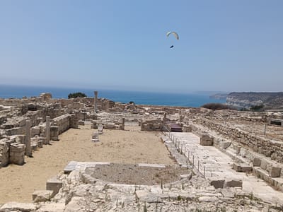 The image captures a scenic view of ancient ruins overlooking a coastal landscape under a clear blue sky. In the foreground, crumbling stone walls and pillars suggest a historical archaeological site, likely an ancient city or settlement. The ground within the ruins is covered in sand and scattered stones, with some paved sections and remnants of foundations visible.

In the mid-ground, the azure sea stretches towards the horizon, meeting a hazy sky. Along the coastline, rocky cliffs and vegetation are discernible. A distinctive element in the sky is a paraglider, with one person harnessed beneath a colorful canopy, soaring high above the water.

The overall scene evokes a sense of history and leisure. The time of day appears to be midday, given the bright sunlight and clear sky. The weather seems warm and pleasant, ideal for outdoor activities like paragliding and exploring historical sites. The location appears to be coastal, with the ruins situated on an elevated position offering panoramic views of the sea. The presence of the paraglider adds a dynamic and adventurous aspect to the otherwise static and ancient landscape. No visible text is present in the image.