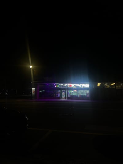 The image presents a nighttime exterior view of "The Corner Smoke & Vape," a commercial establishment located at 3416 SW 34th St, Gainesville, Florida, United States. The scene is dark, illuminated primarily by the bright fluorescent-style lighting from inside the building and a single streetlamp positioned on the left, which creates a lens flare.

The single-story building features a wide glass storefront displaying various products on shelves within the interior. Identifiable signage on the building's facade includes "VAPE" in green neon lettering, "BOBA TEA," and "KRATOM." A larger sign above the main windows reads "THE CORNER SMOKE & VAPE" with additional text indicating "KRATOM & DELTA." Purple neon-like lighting accents the exterior architecture of the building's front.

In the foreground, a paved two-lane road stretches horizontally across the frame. A curb separates the roadway from a partially visible parking area or sidewalk. Faint white parking space lines are discernible on the lower left. A dark-colored vehicle is partially visible, parked on the far left of the frame. To the right of the main building, another structure with illuminated horizontal windows is partially visible. No individuals are present or discernable in the image.