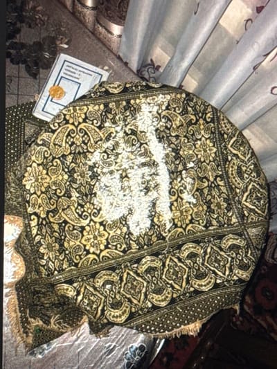 A high-angle photograph captures an indoor scene in Dehqonbozor, Uzbekistan. The central subject is a dark textile, likely a tablecloth or large fabric piece, densely patterned with gold, black, and lighter paisley and floral motifs. A distinct trail of white powdery substance is spread across the central upper portion of this patterned fabric. Behind and slightly to the left of the textile, a light-colored document or booklet is partially visible. Its cover displays Cyrillic text, including "ДЕҲҚОНБОЗОР TУМАНИ" (Dehqonbozor district), and a circular gold-colored seal. The surrounding environment includes a dark, patterned floor or rug with floral designs to the left, and light-colored pleated curtains with subtle reddish-brown patterns to the right. A section of a darker, reddish-toned patterned rug is visible in the bottom right. No individuals are present, and no specific actions or interactions are depicted.