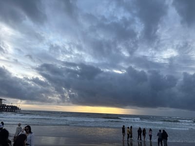 A stormy beach scene under a cloudy sky. People stand near the water's edge, their figures silhouetted. Gentle waves break on the shore, reflecting the dim light. A pier juts into the ocean on the left. The sky is filled with dark, textured clouds, hinting at an approaching storm. A hint of golden sunlight peeks through on the horizon.