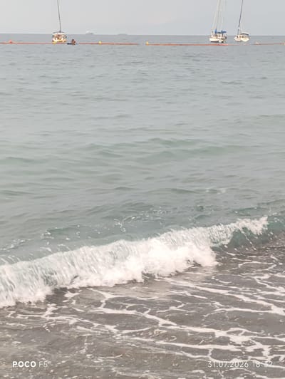 A choppy sea with moderate waves laps at a pebble beach. In the background, several sailboats are visible on the water. A floating orange boom, possibly marking a boundary or containing debris, stretches horizontally across the scene. The sky is overcast. The text "POCO F5" is visible in the lower left corner, and "31.07.2026 18:32" is in the lower right corner, indicating the device and date/time the photo was taken. The location is Camogli, Italy.