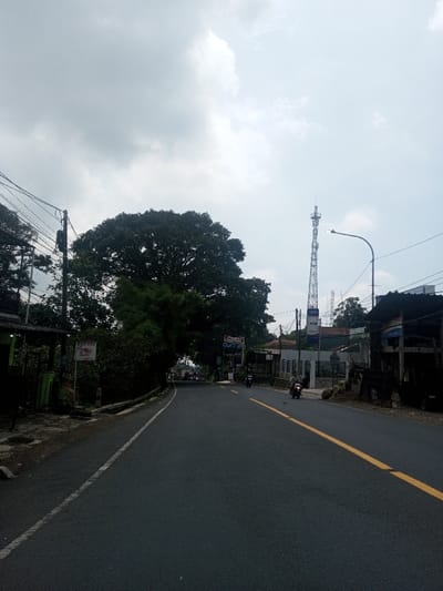 The image displays a roadway in Niz Lakuk, India, viewed from a low angle under an overcast sky with grey clouds. The dark asphalt road features a solid white line along its left edge and a broken yellow centerline, suggesting two-way traffic. The road curves gently to the left in the distance.

On the left side, dense green foliage and a large, dark-canopied tree extensively overhang the road. A utility pole with a dense network of black wires runs along this side, where a building with green accents and a white rectangular sign displaying red text (partially visible, possibly "RASA") is located.

On the right side of the road, further into the mid-ground, a tall, grey metal communications tower equipped with multiple antenna arrays is a prominent feature. Below the tower, commercial structures are visible, one featuring blue and white "ATM" signage. A street light pole with a curved arm and an illuminated fixture is positioned in front of these buildings. Multiple motorcycles are present on the road: one with a rider in a light-colored shirt is traveling away from the viewer in the right lane, and another motorcycle with a rider is further down the road, also moving away. Additional indistinct vehicles or figures are present in the distance under the large tree. The immediate right foreground includes dark, rustic-looking structures, some with corrugated metal roofs.