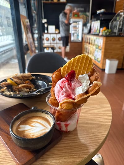 This is a close-up shot of a dessert and coffee on a wooden table in what appears to be a cafe. In the foreground, a black ceramic cup with latte art sits on a dark wooden coaster. To the right, a bubble waffle cone filled with pink ice cream, whipped cream, and a strawberry is prominently displayed in a clear plastic cup. A partially eaten plate of food, possibly fried chicken or seafood with lemon, is visible in the background on the left. The cafe's interior is blurred, showing shelves with food items, a counter, and a person in the distance. The overall impression is a casual dining scene. The text overlays suggest that this image was taken in Tha Chana, Thailand.