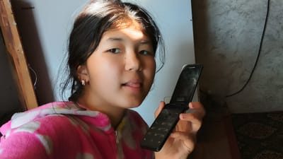 A young woman with dark hair, wearing a pink and white patterned robe, is holding a black flip phone in her right hand, positioned to the right of her face. Her tongue is sticking out slightly. The background includes a white surface with the word "Hisense" visible in the upper left corner, and a textured grey wall on the right. A wooden frame is to the left of the woman. The scene appears to be indoors, in what could be a residential setting in Uchkun, Kyrgyzstan, given the context.