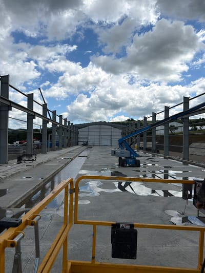 This is a full, eye-level, outdoor shot of a construction site in Stróża, Poland. The image captures a large, partially constructed steel building with a light-colored, curved roof. A blue boom lift is positioned in the middle ground, with its arm extended upwards, resting on one of the building's steel beams. The ground is concrete, with puddles of water reflecting the sky and clouds. In the foreground, a yellow metal safety railing is visible, partially obscuring the view. The sky is filled with dramatic, white and gray cumulus clouds against a bright blue backdrop. In the distance, green hills and trees are visible. The scene appears to be a typical construction project underway on a cloudy day.