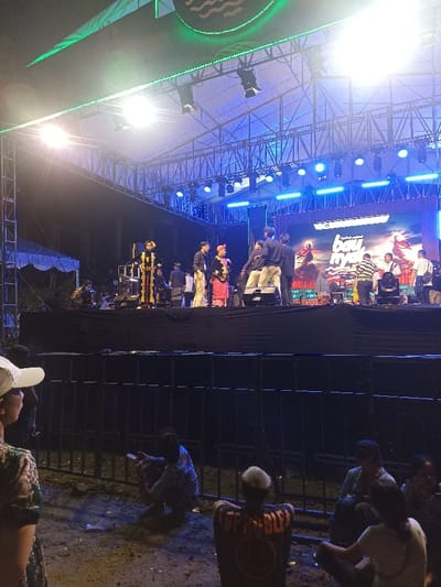 This image depicts an outdoor night event centered around a large, brightly lit stage in Kecamatan Pujut, Indonesia. The stage is a robust structure built with metal trussing, supporting numerous blue and white stage lights, as well as green accent lighting along its upper frame. At the very top, a banner or screen displays a circular logo with wavy lines.
On the stage, several people are visible. Two individuals stand out due to their traditional attire: one on the left wears an elaborate dark costume with gold embellishments and a tall headdress, while another near the center is dressed in reddish-pink traditional garments and headwear. Other individuals in casual clothing are also present on the stage, some seemingly involved in preparations or as part of the event crew. Behind the performers, a large LED screen displays dynamic, colorful graphics with visible text, including parts of words like "bau..." and "NYANYI," indicating a performance or cultural show.
In the foreground, a metal barrier separates the stage from the audience area. Below this barrier, several spectators are sitting directly on the dirt ground. One person is engrossed with their mobile phone, while another, with short blonde hair, wears a black t-shirt featuring a prominent circular logo on the back. A person wearing a white cap is partially visible on the far left. The scene is illuminated almost entirely by the stage lights, creating a bright focal point against the dark night sky, suggesting an evening or nighttime cultural performance or concert.