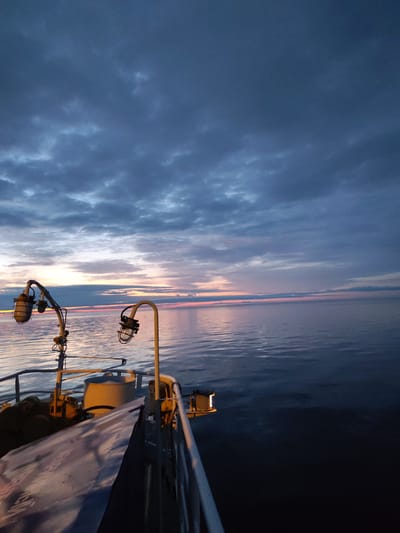 The image depicts a view from the deck of a vessel at sea, likely near Shcheleiki, Russia, at twilight. The sky is filled with dramatic, dark clouds, with a sliver of a pink and orange sunset visible on the horizon. The water is calm, reflecting the muted colors of the sky. On the deck of the boat, two industrial-style lamps on yellow metal arms are visible, casting a warm glow. Part of the boat's railing and a white tarp-covered object are also in the foreground. The overall impression is one of a serene but potentially moody maritime scene.