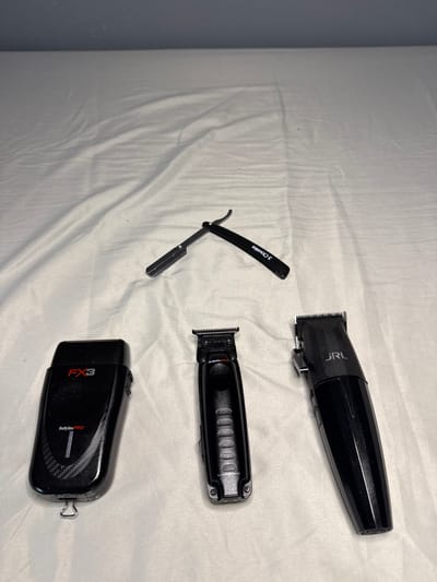 The image shows four black grooming tools laid on a white cloth surface. The tools include a straight razor, a hair clipper with "JRL" printed on it, another hair clipper with multiple holes on its body, and a foil shaver labeled "FX3" and "BabylissPRO". The setting appears to be indoors, with a gray wall visible on the left edge of the frame. The photograph does not provide any information to confirm if the location is in Edgartown, United States of America.